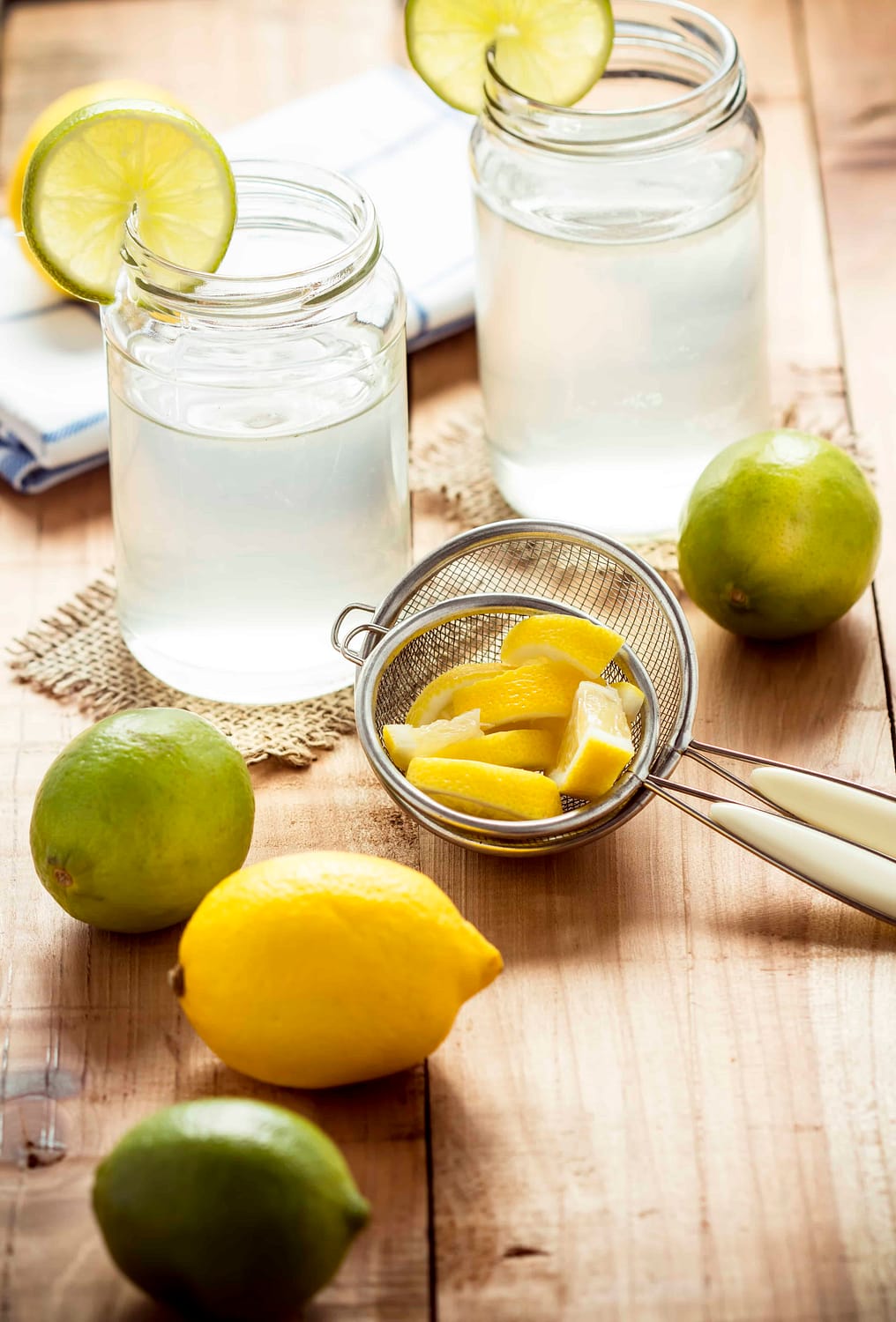 Lemons and limes on a wooden table next to a juice strainer and a glass of lemonade