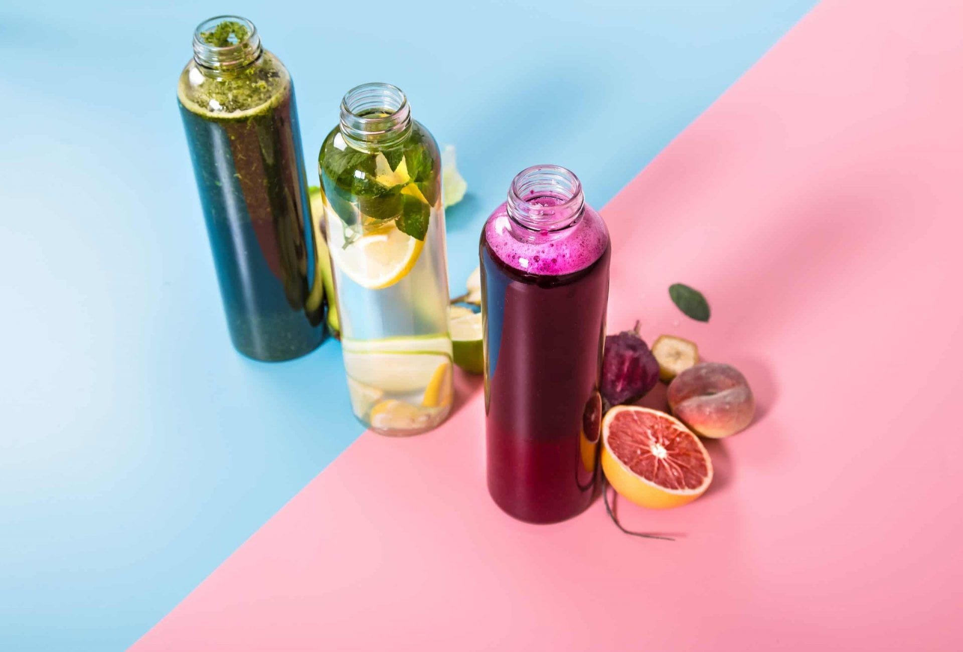 Glass juicing bottles with natural detox drinks and a variety of fruits and vegetables on a pink and blue background .