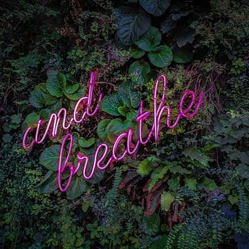 image of a pink neon sign with words: "and breathe" against a garden background