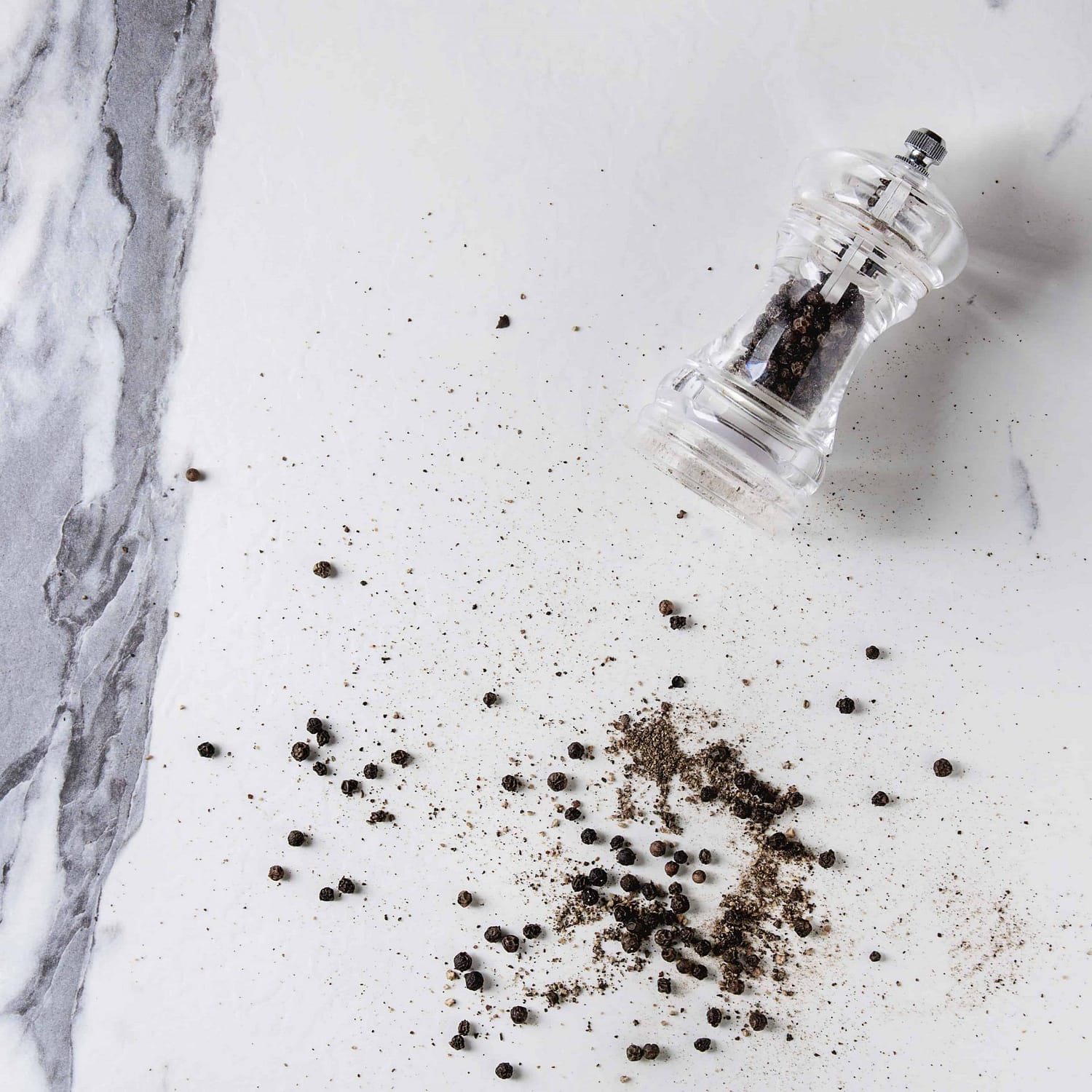 Black peppers peppercorns and ground powder from transparent pepper mill over white marble 