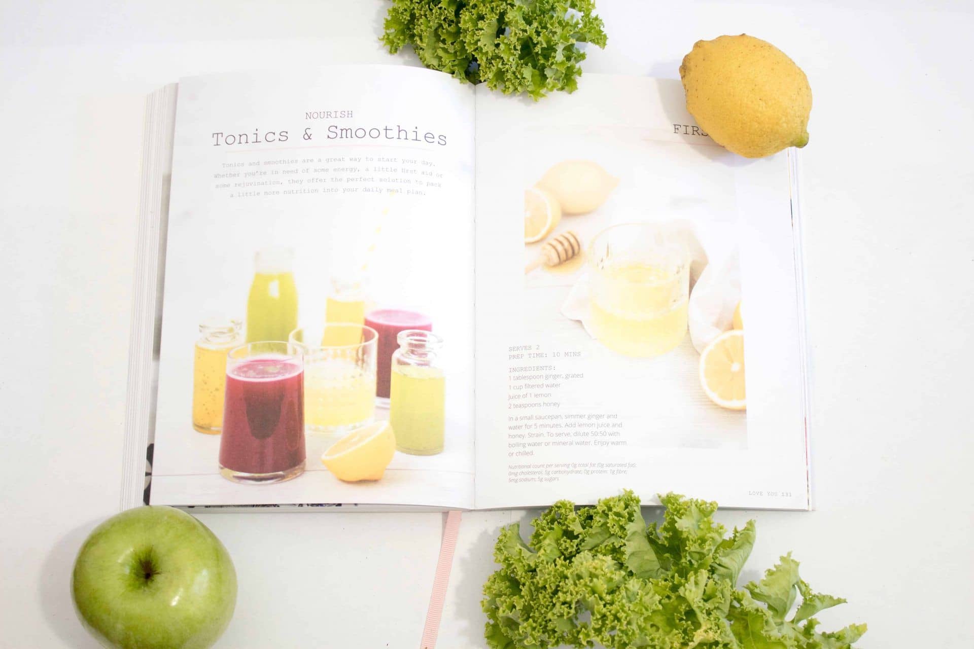 Open vegan smoothie cookbook with ingredients kale, lemon, and apple sitting next book