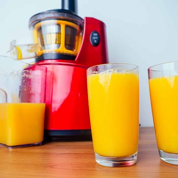 Image of a red masticating juicer on a table next to 2 glasses of orange juice