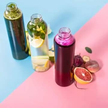 Glass juicing bottles with natural detox drinks and a variety of fruits and vegetables on a pink and blue background .