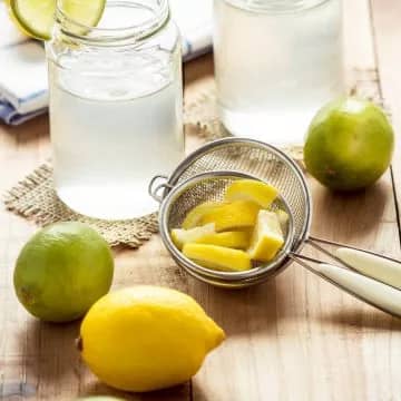 Lemons and limes on a wooden table next to a juice strainer and a glass of lemonade